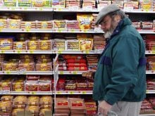 A bearded middle-aged man wearing glasses and a tam-style hat picks up a package of wieners in a WalMart grocery store.