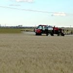 A high clearance sprayer sprays a crop with a concrete inland grain terminal visible in the background.
