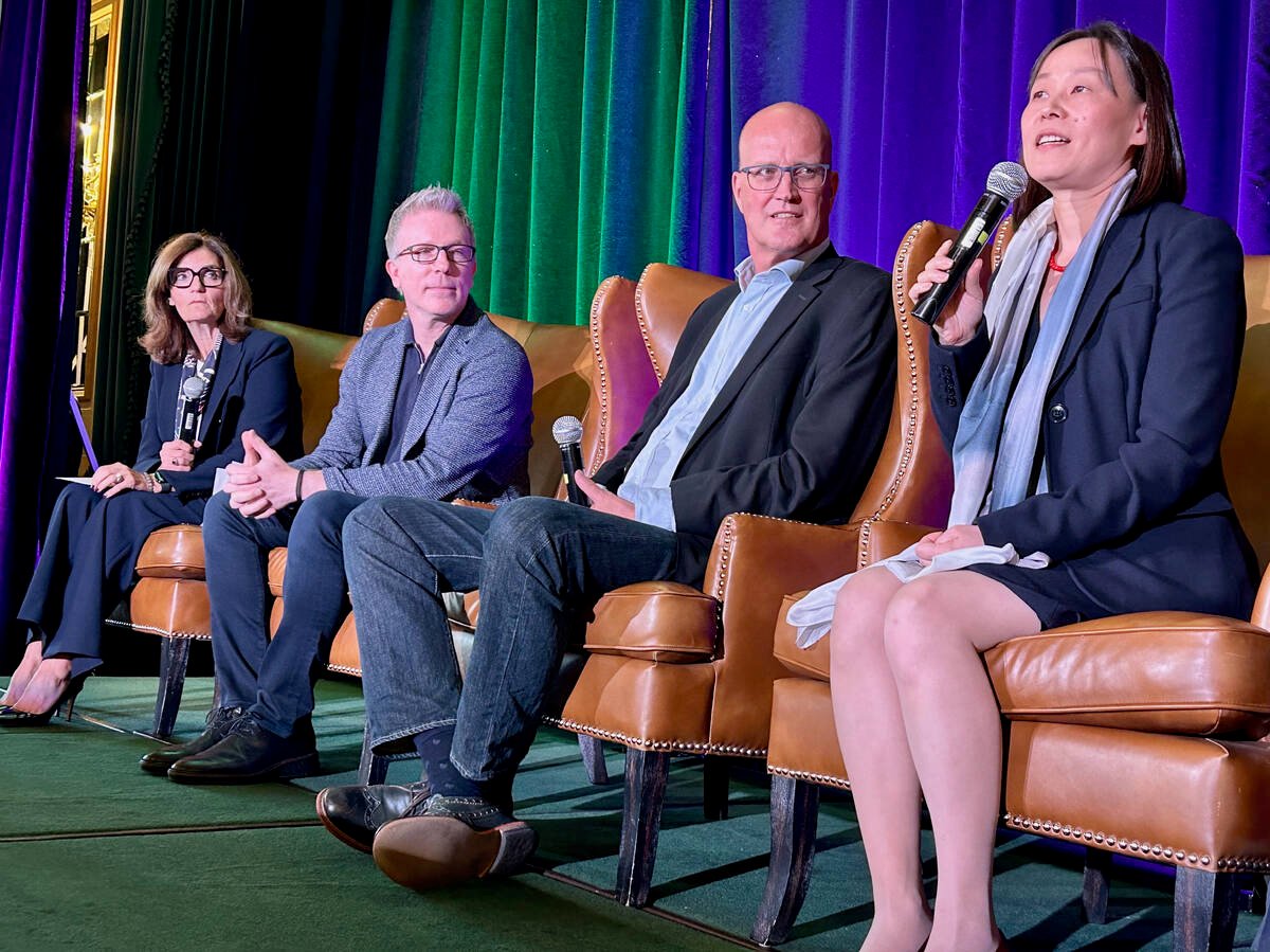 Four people, two women and two men, sit on comfortable brown leather wing-back chairs arranged in a straight line beside one another on a stage during a conference. Each has a microphone in their hands and the woman closest to the camera is speaking into it.