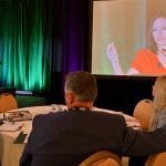 Two people seated at a round table near the stage at a conference look toward a woman speaking at a podium as her image is simultaneously projected onto a large screen behind her for those further away to see.