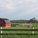 A pumpjack works in a field behind an old red barn with some weathered gray wooden corrals beside it.
