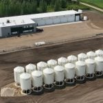 An aerial view of the Humaterra plant near Birch Hills, Saskatchewan, showing a large rectangular shop and 20 white upright smooth-walled hopper bins neatly positioned in two rows of 10.