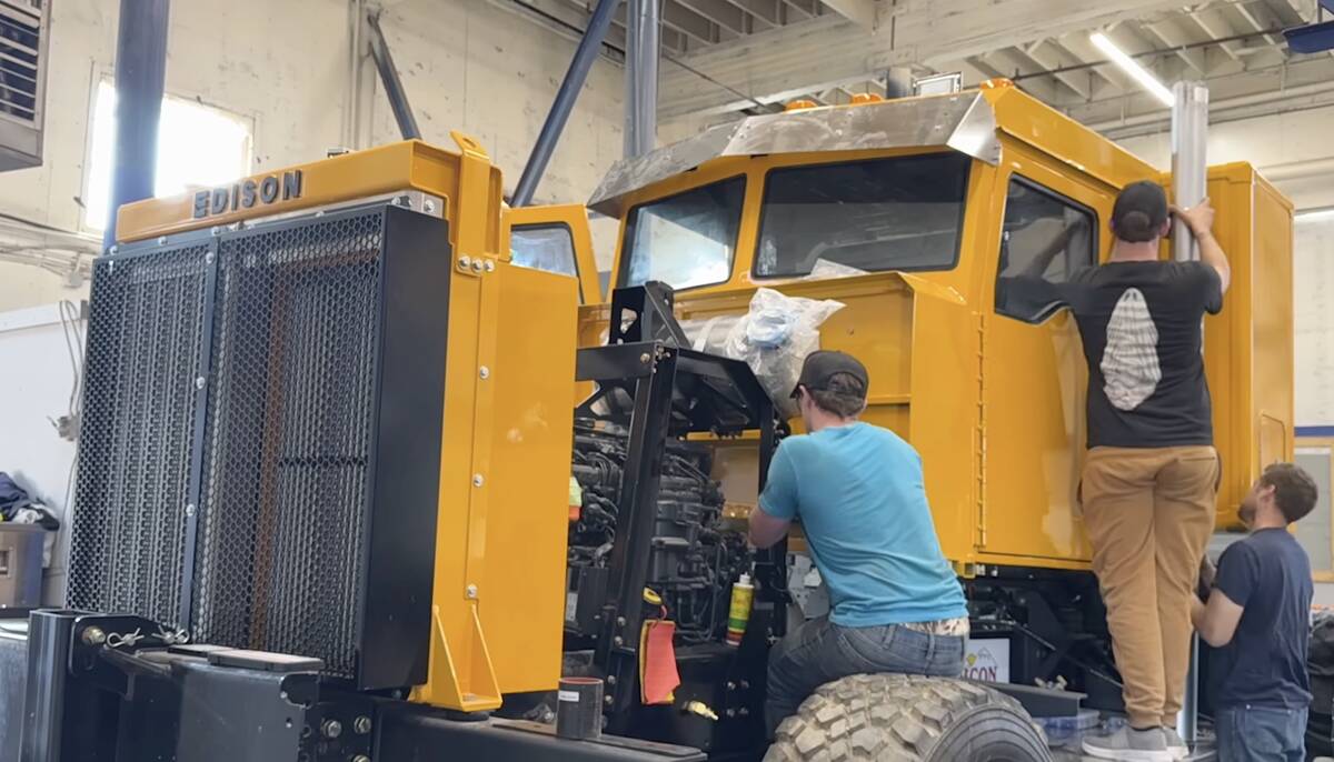 Three men working on a large yellow truck with an "Edison" badge on the front and the side panels removed in a shop.