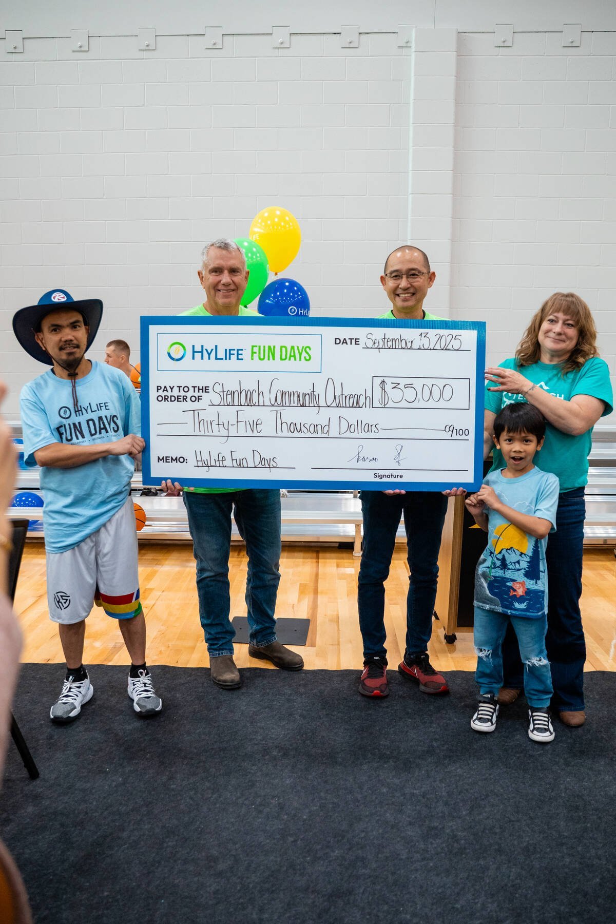 Four adults and a child stand in a gym holding a large cheque for $35,000 from HyLife Fun Days payable to "Steinbach Community Outreach."