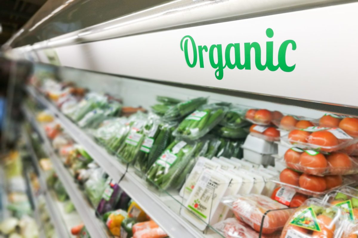 A close-up of the green "Organic" script on a white background labelling a section of a cooler in a grocery store, with packaged tomatoes and other types of organic produce on the shelves beneath it.
