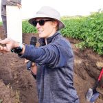 A woman standing in a pit about four feet deep dug in a field wearing a hat, sunglasses and holding a microphone offers a handful of soil to an unseen person for them to examine.
