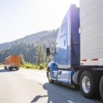 Two semi trucks go around a curve on a highway in a mountainous area on a sunny day in the summer.