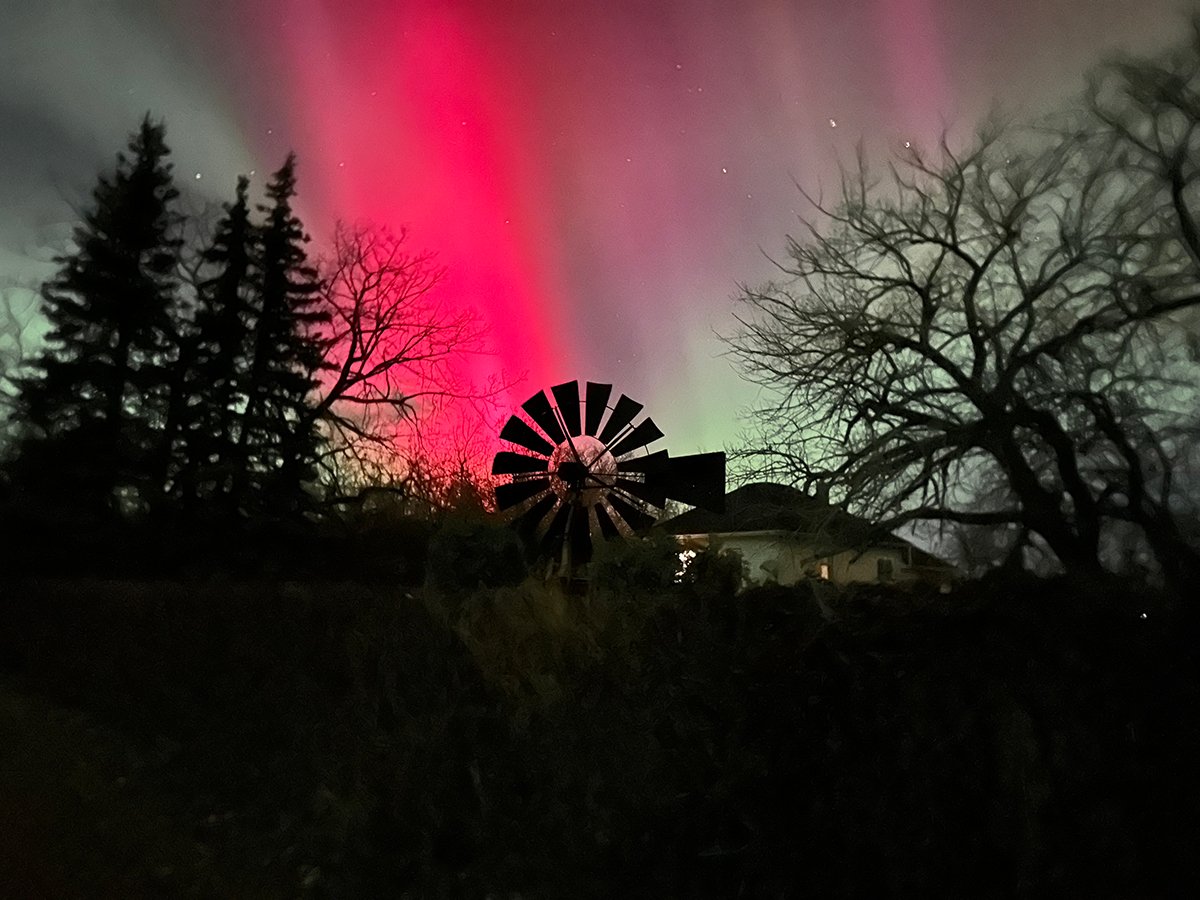 A brilliant red "stripe" of aurora lights up the sky over an old fashioned windmill with a house visible in the background.