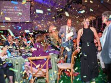 Three people, a man with crutches between two women, walk between tables with peope seated at them while confetti fills the air at an awards gala.