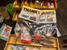 A yellow fishing tackle box sits open on a table with a number of lures in plastic bags on top with a label that reads, "Thanks for hooking up with us."