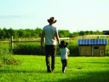 A farmer and his young son, both wearing cowboy hats, hold hands as they walk away from the camera on green grass toward a small shed in the distance.