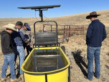 Four farmers stand around a yellow stock water tank in a pasture. The tank has a solar panel attached above it for power.