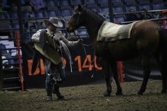 A man wearing a cowboy hat and jeans stands on the dirt floor of an arena holding a horse's reins with his left hand as he prepares to throw a saddle onto the blanket on the horse's back as he competes in a "Ranch Rodeo" timed event at Canadiqn Western Agribition in 2025.