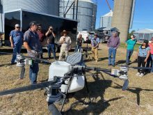 A large "quad copter" style drone sits on the ground as Adrian Rivard talks to attendees of a cover crop seeding demonstration in the background.