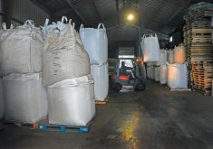 Bulk bags are stacked two-high in a warehouse while a forklift operates in the aisle between them.