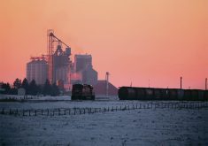A CP train engine moves toward some grain cars on a siding near an inland terminal visible in the background near sunset in the winter.
