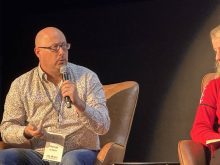 A bald man is sitting in a chair on stage holding a microphone.