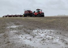 A low angle photo of a puddle on top of the soil in a field with a large, tracked tractor hitched to an air seeder parked in the background on a rainy day.