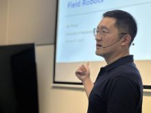 Jay Wang, assistant professor from the University of Manitoba faculty of Mechanical Engineering, stands in front of a screen speaking to an unseen group.