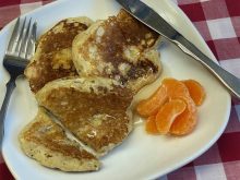 Barley pancakes with syrup on top sit on a white plate with a knife and fork and few grapefruit segments on the side.