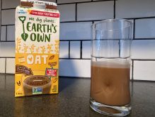 A low angle photo of an "Earth's Own" chocolate oat milk container on a countertop next to a clear glass slightly less than half full of the beverage.