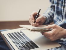 The hands of a man are holding a notebook and writing in it overtop a laptop computer sitting on a desk.