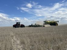A large, tracked tractor pulling an air seeder rig crosses from right to left in front of the camera on a sunny day with a few puffy white clouds in the sky.