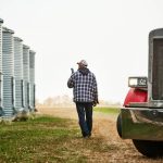A man in a ball cap and blue plaid chore coat walks along beside some steel grain bins while talking on his smartphone.