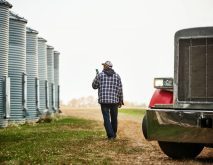 A man in a ball cap and blue plaid chore coat walks along beside some steel grain bins while talking on his smartphone.