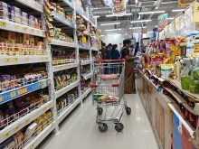A grocery cart is parked in the middle of an aisle in a supermarket in Mumbai, India, while a woman looks at a product behind it.