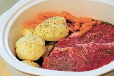 A raw piece of meat sits in a white baking dish with whole, peeled potatoes and carrots beside it - all sprinkled with seasoning, ready to put into the oven.