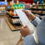 A close-up of a long paper grocery receipt being held by a woman in a grocery store.
