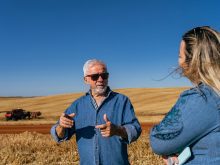 A female journalist with her back to the camera talks to a farmer at harvest in the middle of a field under a clear blue sky. The farmer's combine is parked in the background.