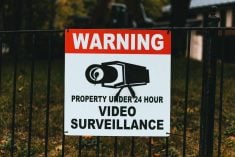A "WARNING - Property Under 24 Hour Video Surveillance" sign is zip tied at all four corners to a black, wrought iron fence.