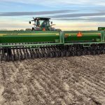 A farmer looks over his shoulder at a green John Deere seeder as he rolls along seeding a demonstration plot.