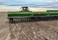 A farmer looks over his shoulder at a green John Deere seeder as he rolls along seeding a demonstration plot.