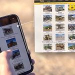 A man's hand holds a smartphone scrolling with his left thumb over items up for auction on an auction website. The same website is seen on a laptop computer to the right of the smart phone.