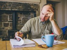 A man sits at a table with with one hand on his forehead and the other leafing through a pile of paperwork.