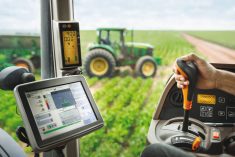 A farmer's hand is seen on a joystick in the cab of a modern tractor with a computer screen on the left and another tractor visible through the cab's glass windows in a field nearby.