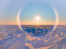 Ice crystals form a halo around the sun that sits low in the sky over a frozen, snowscape below.