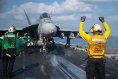A sailor in a yellow top wearing a white helmet holds both arms upward in front of an F18 fighter jet about to be launched from an aircraft carrier during the conflict in Iran in March, 2026.