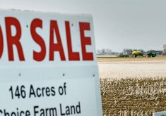 Close-up of a "For Sale 146 acres of choice farm land" sign with an air seeder rig operating on a stubble-filled field in the background.