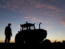 A farmer stands next to a tractor in a field, both perfectly silhouetted against the rising sun.