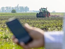 A man's hand holds a remote control, slightly blurred, in the foreground while a tractor pulling a piece of tillage equipment in a field is clear in the background.