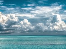 Clouds are seen at various levels overtop a turquoise, tropical ocean.
