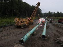 A tracked crane lifts a section of liquid natural gas pipeline under construction on a muddy dirt path next to some trees.