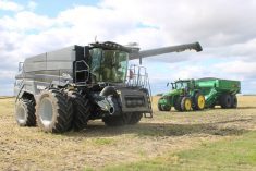 A Fendt combine sits in a field while a a John Deere tractor pulling a grain wagon approaches.