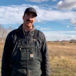 A man with a mustache and a ball cap and wearing black Carhart coveralls stands in a pasture with some black cattle grazing in the distance behind him.