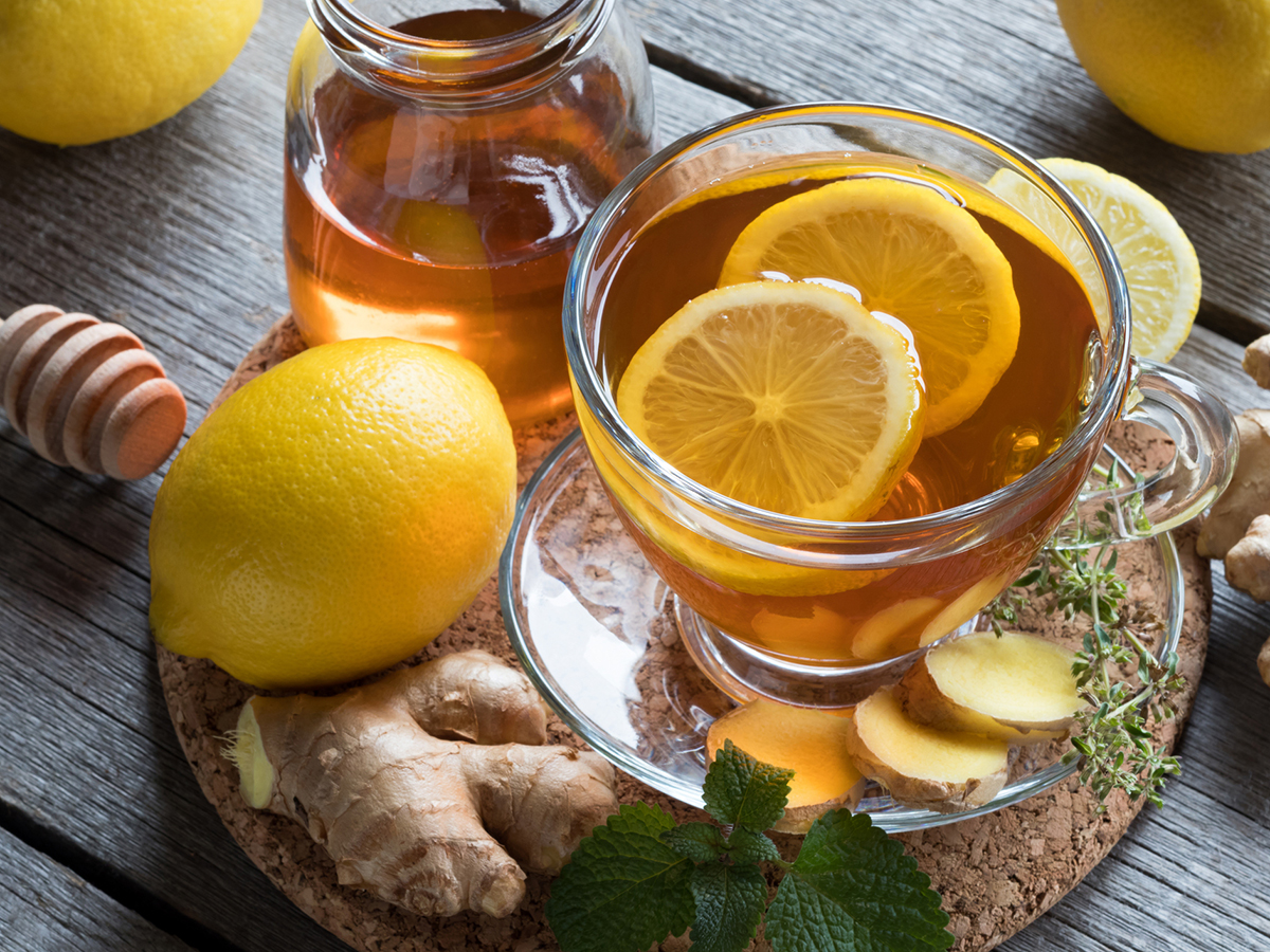 A cup of tea with two lemon slice in it sits on a cork hot plate with some honey and ginger root next to it.