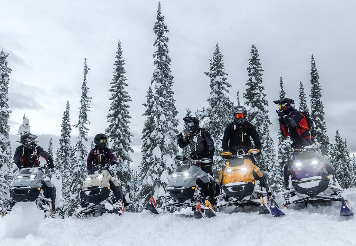 A low angle photo of five sleds and their riders atop a small snowy hill with pine trees in the background.